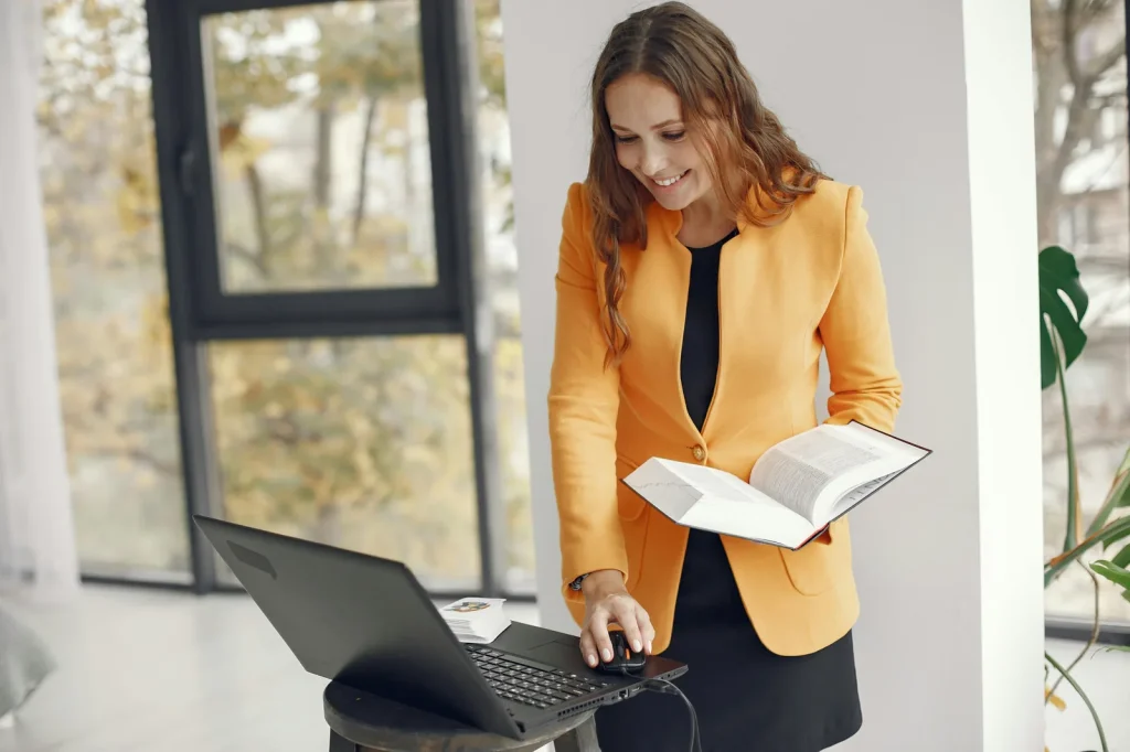 Professional woman reading AI cybersecurity book with laptop in bright modern office representing artificial intelligence in cyber security books reading list