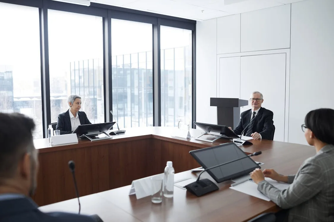 Formal board meeting room with professionals seated at advisory table with microphones and monitors