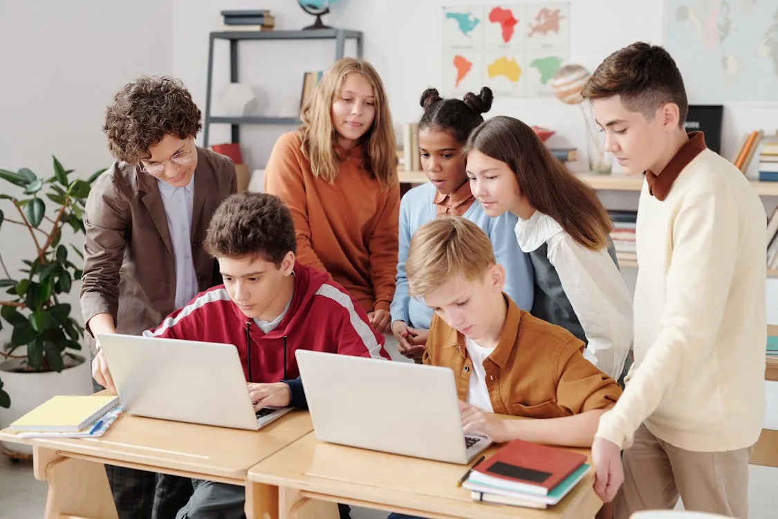 Diverse group of students working on laptops in a classroom with teacher guidance representing school network and data security in K-12 cybersecurity programs