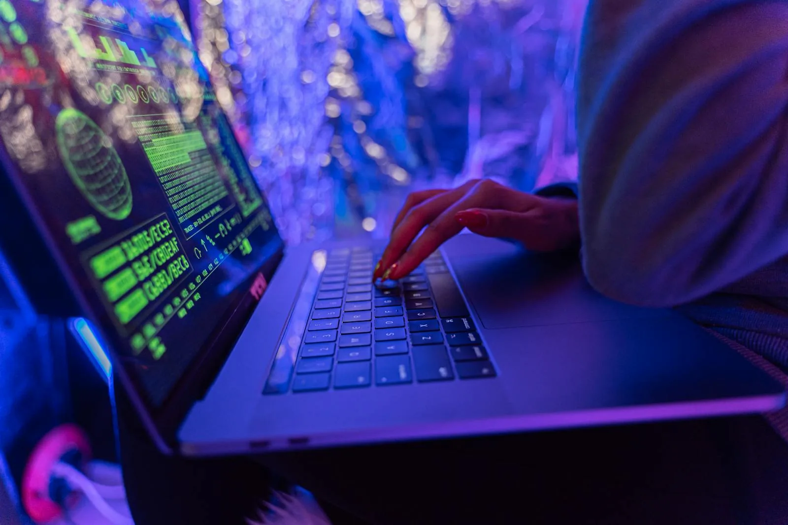 Hands typing on laptop displaying green security code and threat data in neon-lit environment