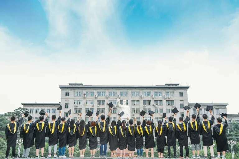 Group of university graduates in black gowns and yellow stoles holding up graduation caps at commencement ceremony