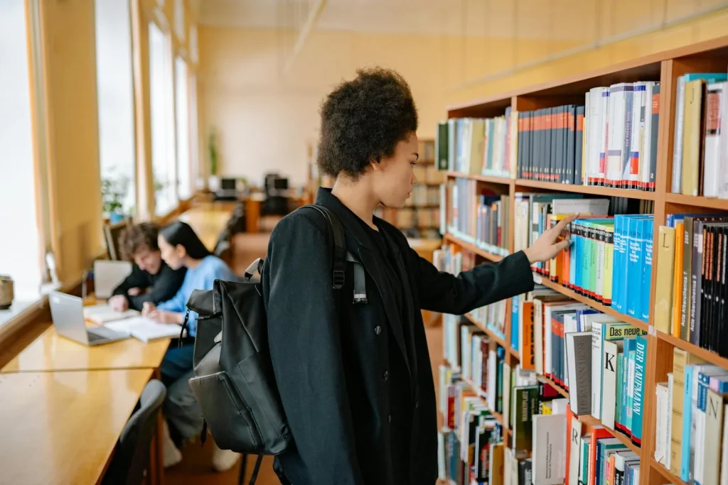 Graduate student with backpack selecting a book from a university library bookshelf with other students studying in background