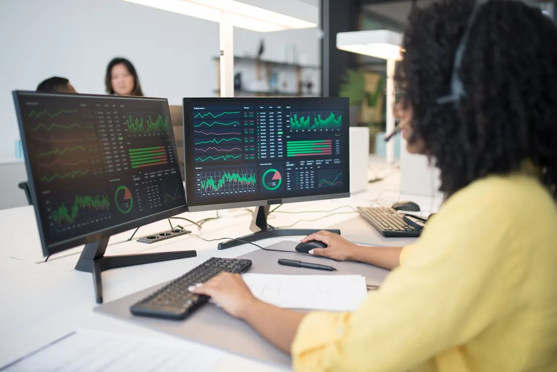 Black woman analyst with headset at dual monitors showing analytics dashboards in bright white office representing SOC alert fatigue and security intelligence operations challenges