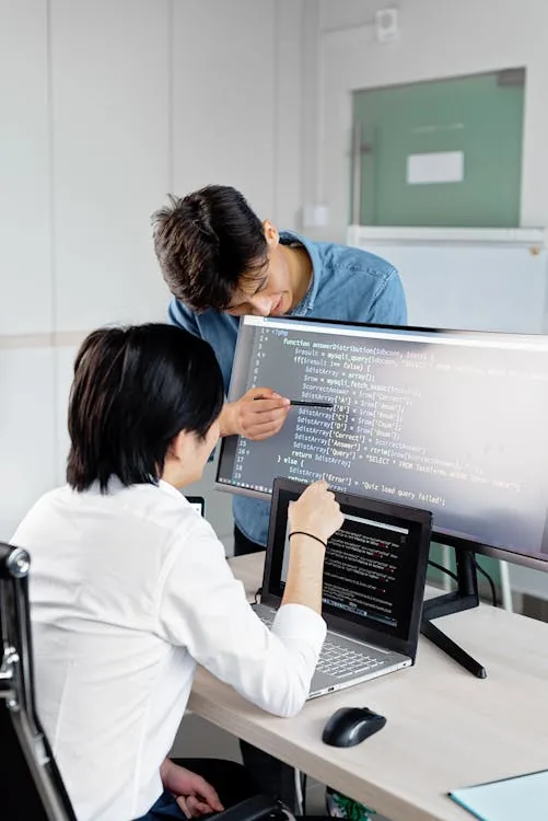 Two software developers reviewing PHP code on large monitor and laptop for application security vulnerability testing